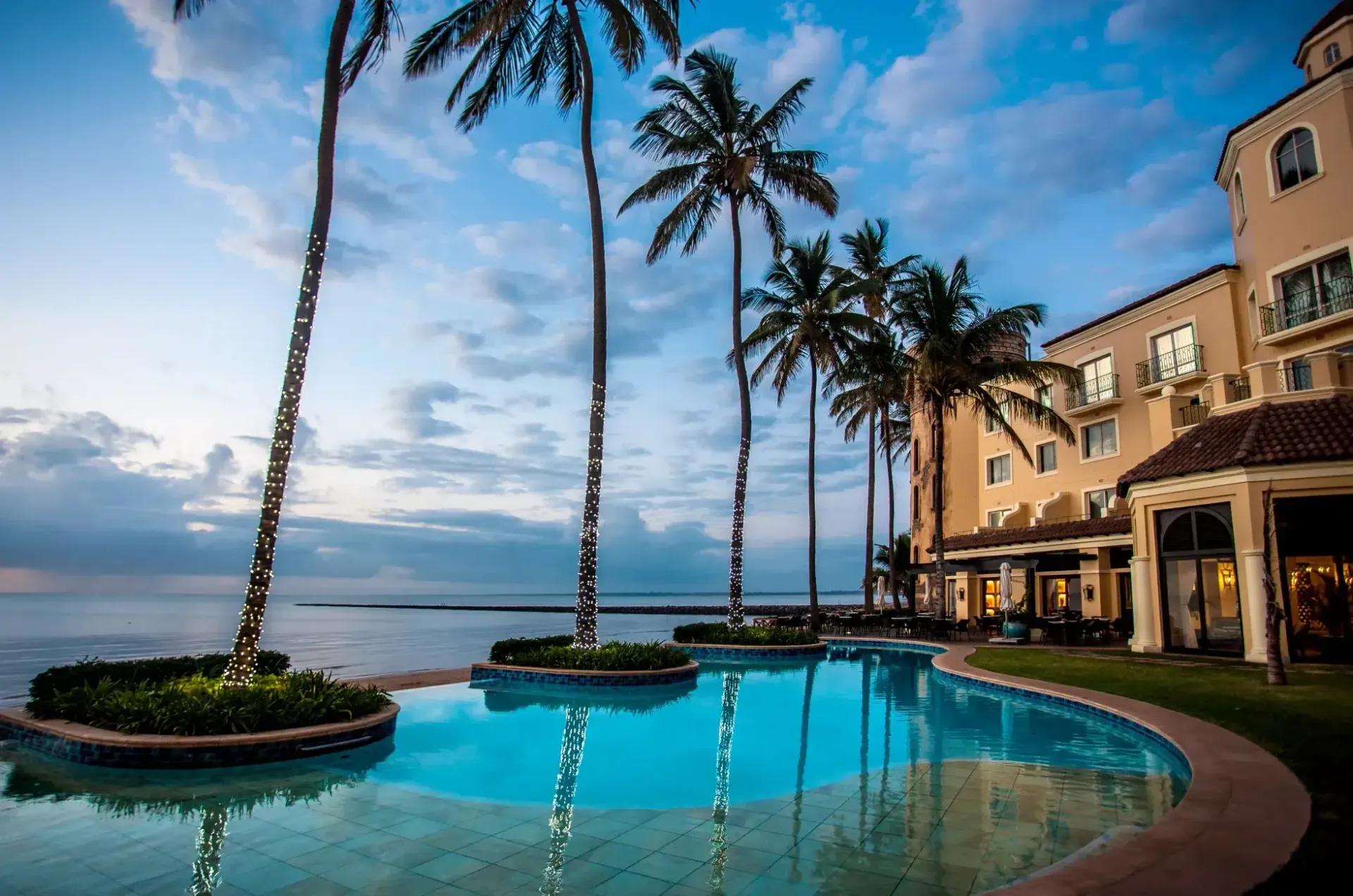 Palm-lined resort pool at sunset overlooking the ocean and sky.