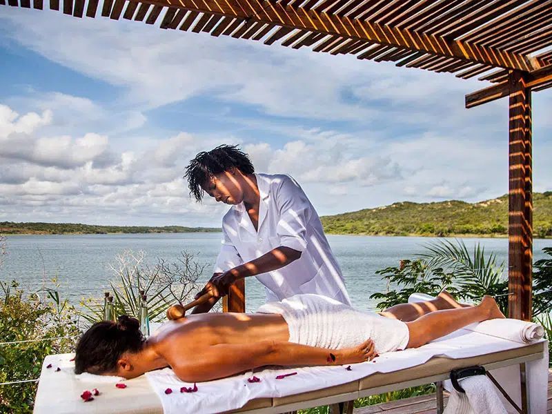 A person gives a back massage with a wooden tool to a client on a table outdoors by a lake under a wooden structure.