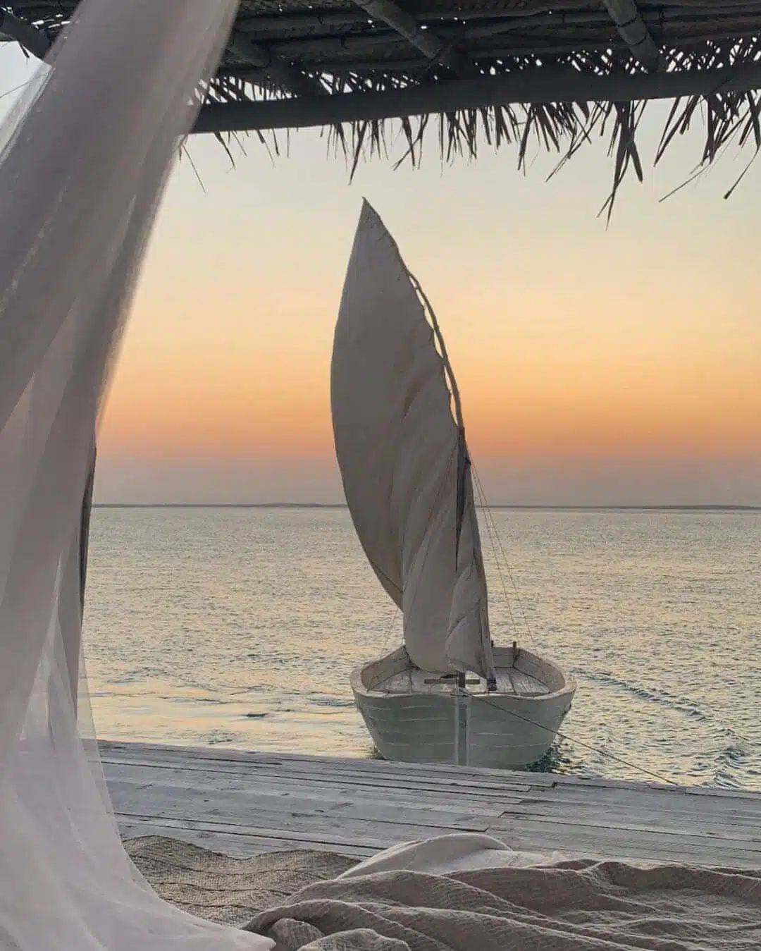 Sailboat at sunset on calm sea, framed by a beach cabana