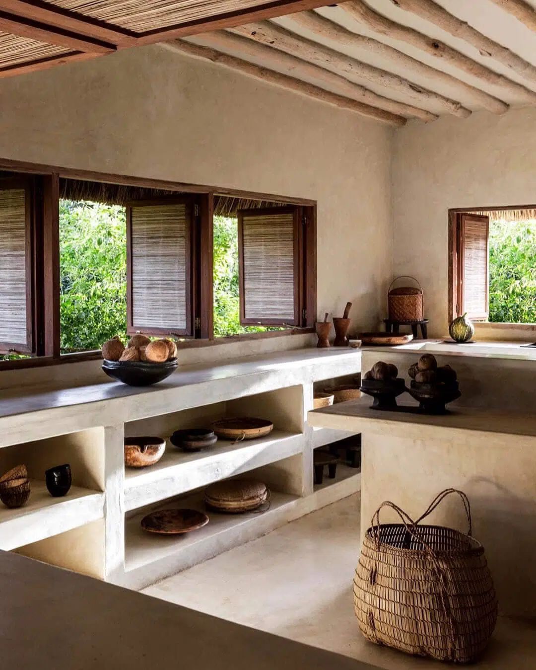 Rustic kitchen with open shelves, clay pots, woven basket, and windows overlooking greenery