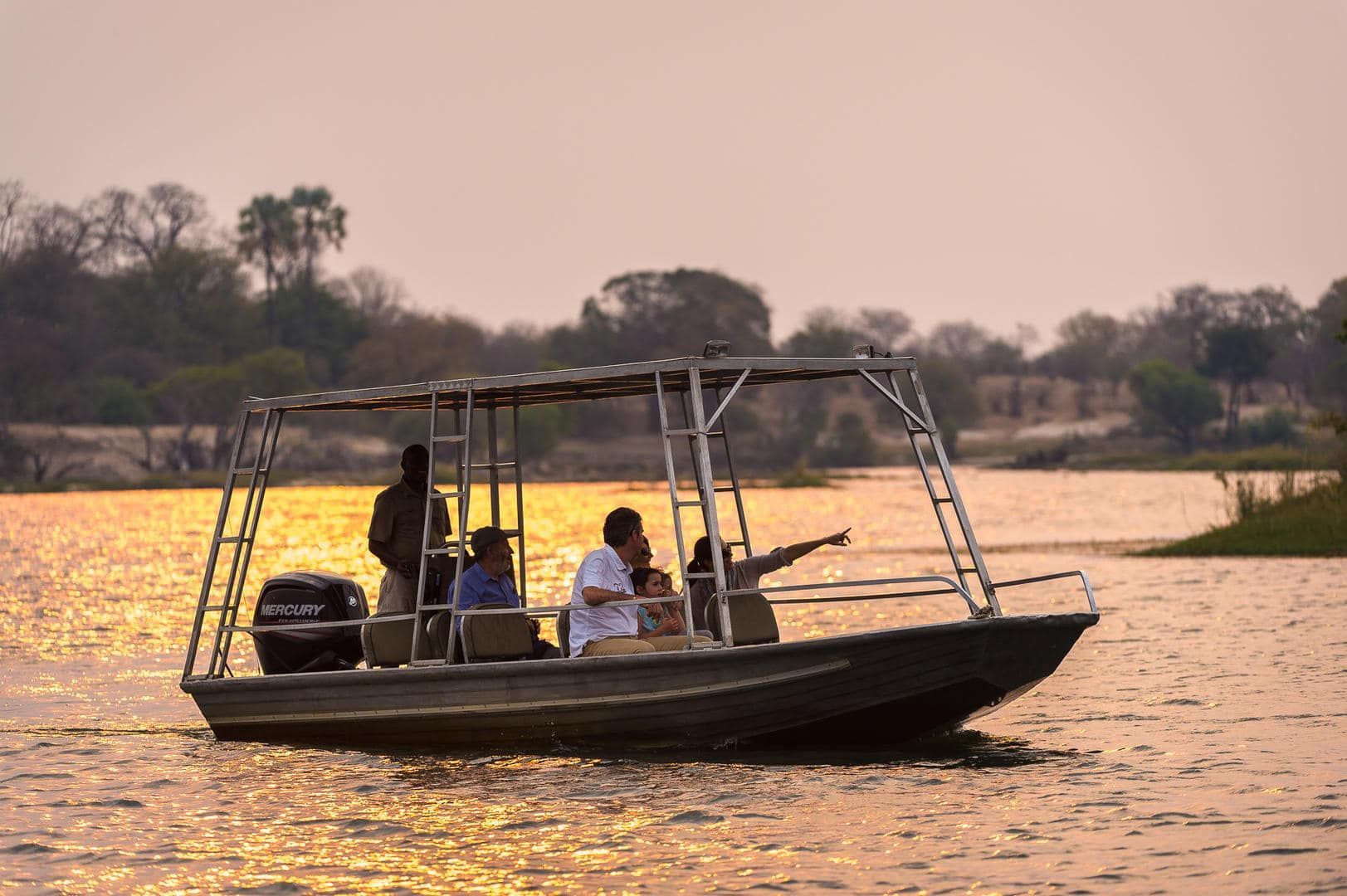 Boat with people on a river at sunset, with golden water and trees along the shore.