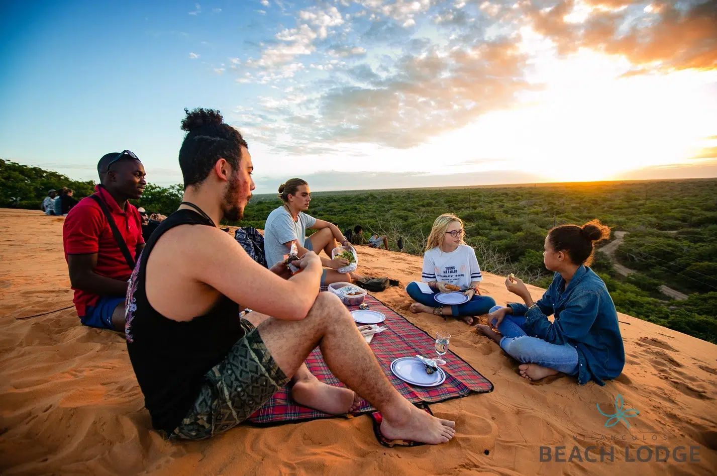 Group sitting on blankets outdoors at sunset, chatting on a scenic hillside with warm orange sky.