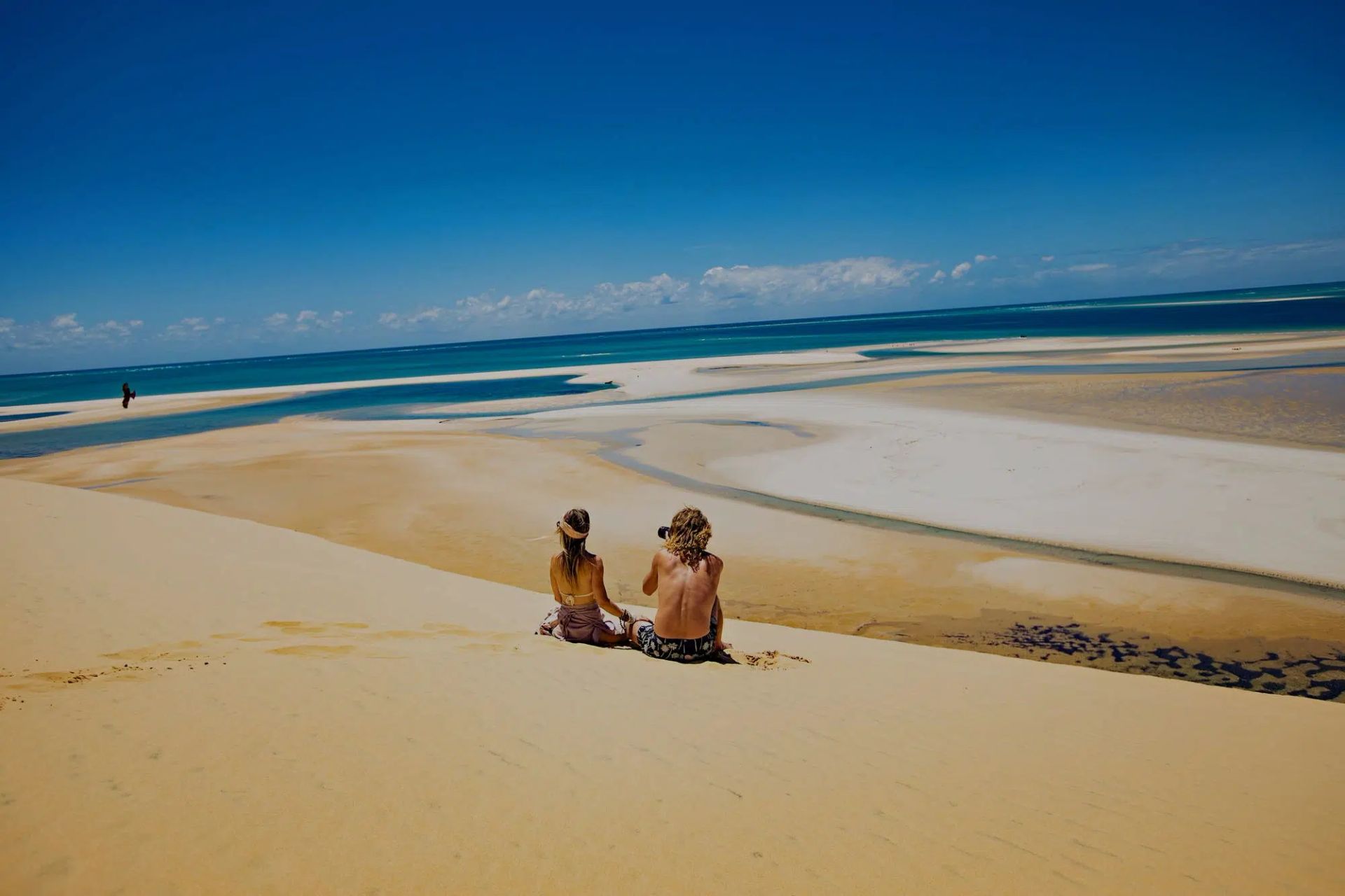 Two people sitting on a sandy beach overlooking blue water and a bright sky