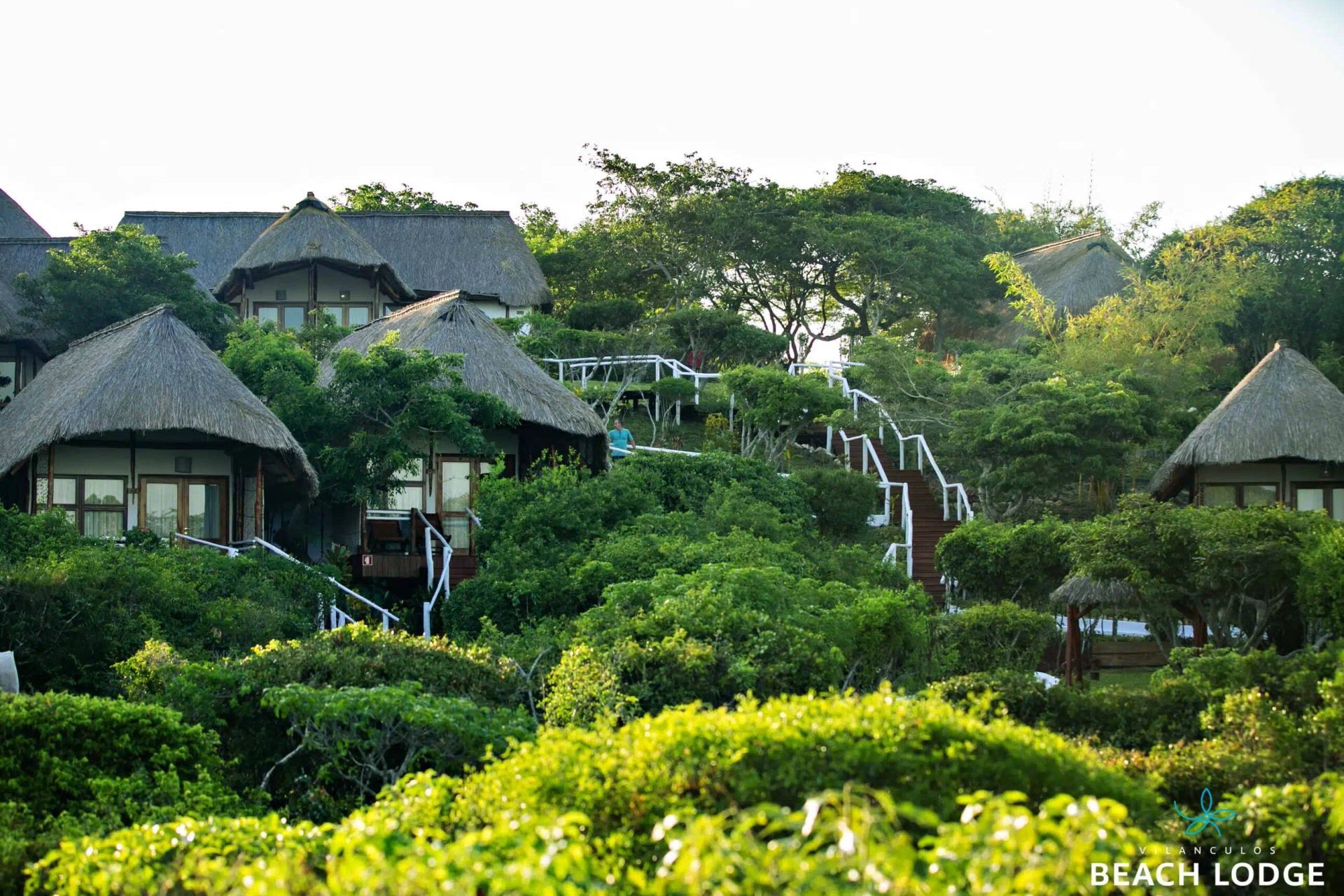 Lush green hillside resort with thatched-roof cottages and winding white walkways