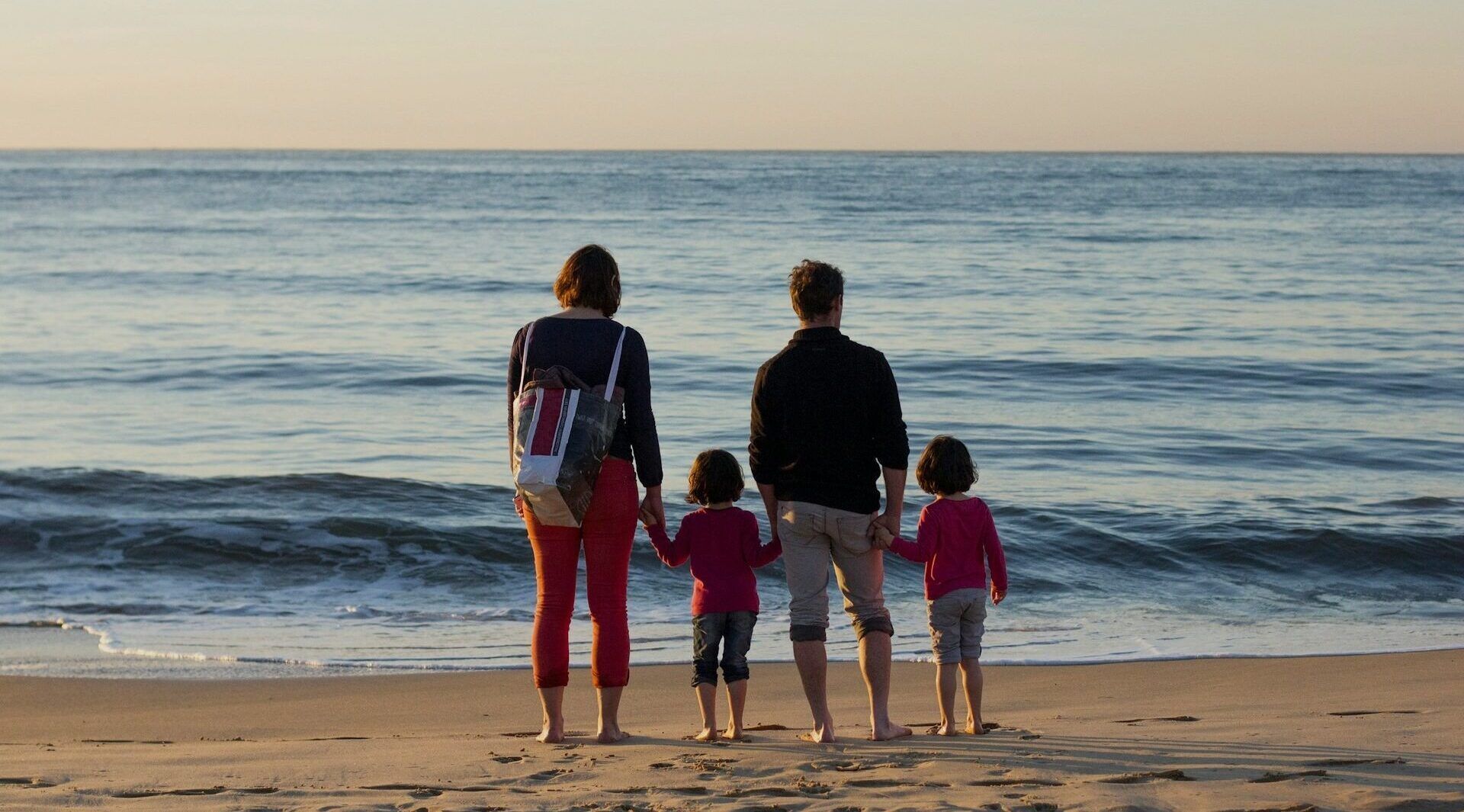 Four people stand on a sandy beach facing the ocean at sunset.