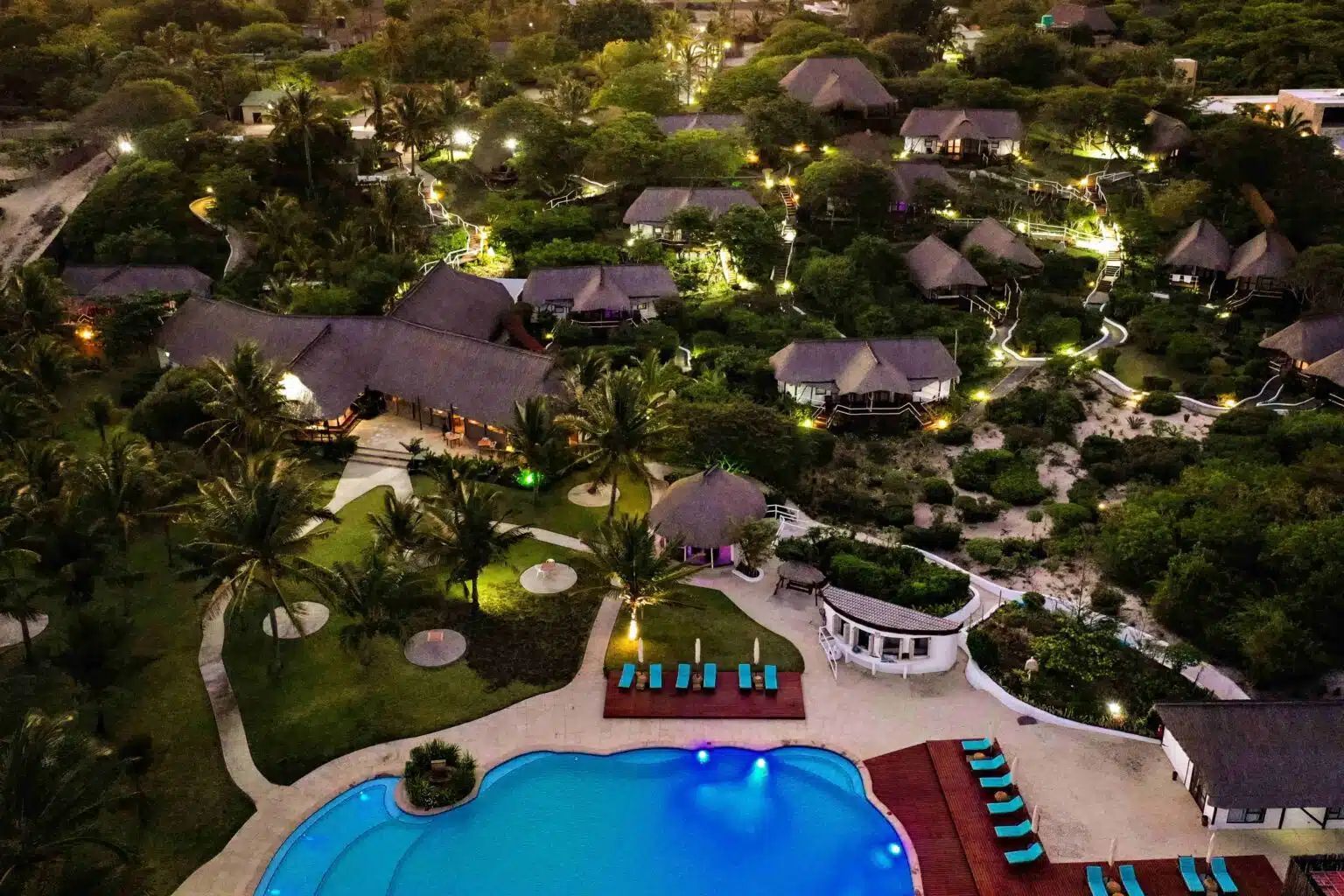 An aerial view of a tropical resort at dusk, featuring a blue swimming pool and multiple thatched-roof villas.