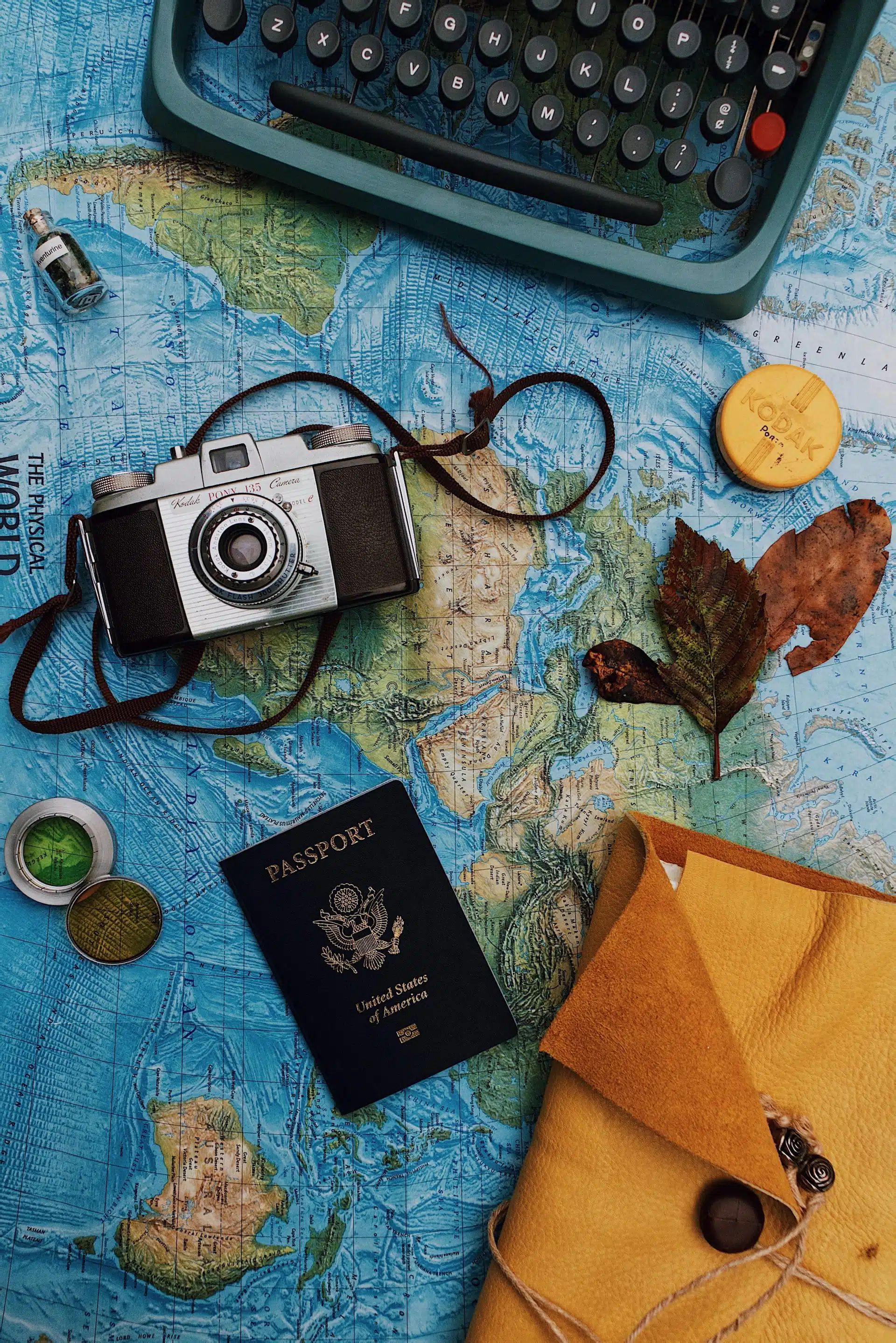 Vintage travel flat lay on a blue map: camera, passport, typewriter, leaves, and a yellow envelope.