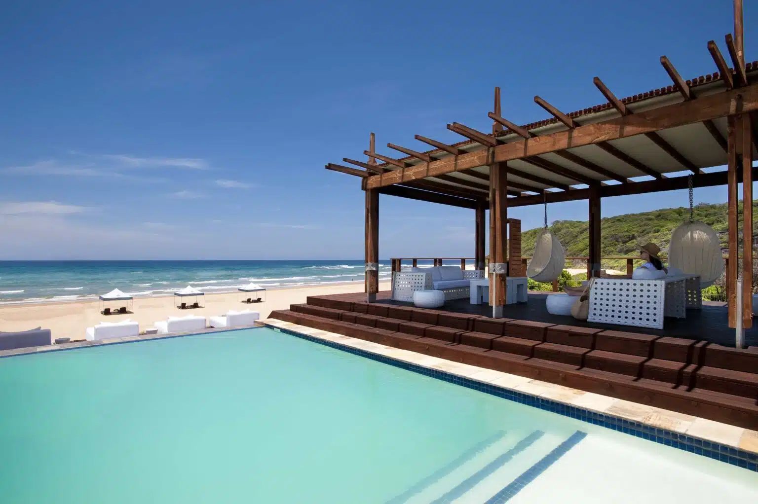 A turquoise infinity pool beside a beach lounge with a wooden pergola, white seating, and views of the blue ocean.