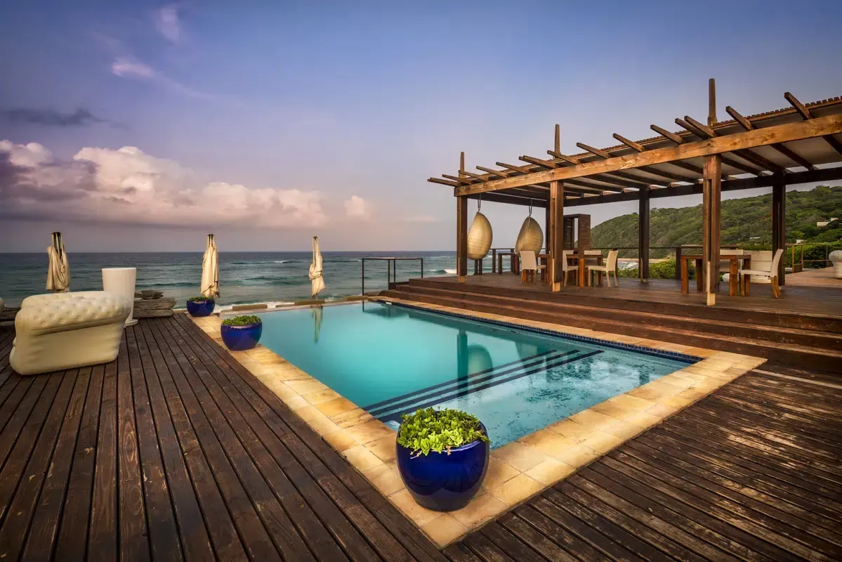 Oceanfront pool and wooden deck with lounge chairs at sunset under a pergola