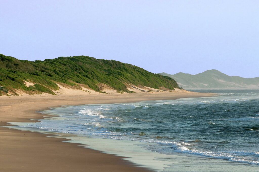 Sandy beach with waves, green dunes, and distant mountains under a clear sky