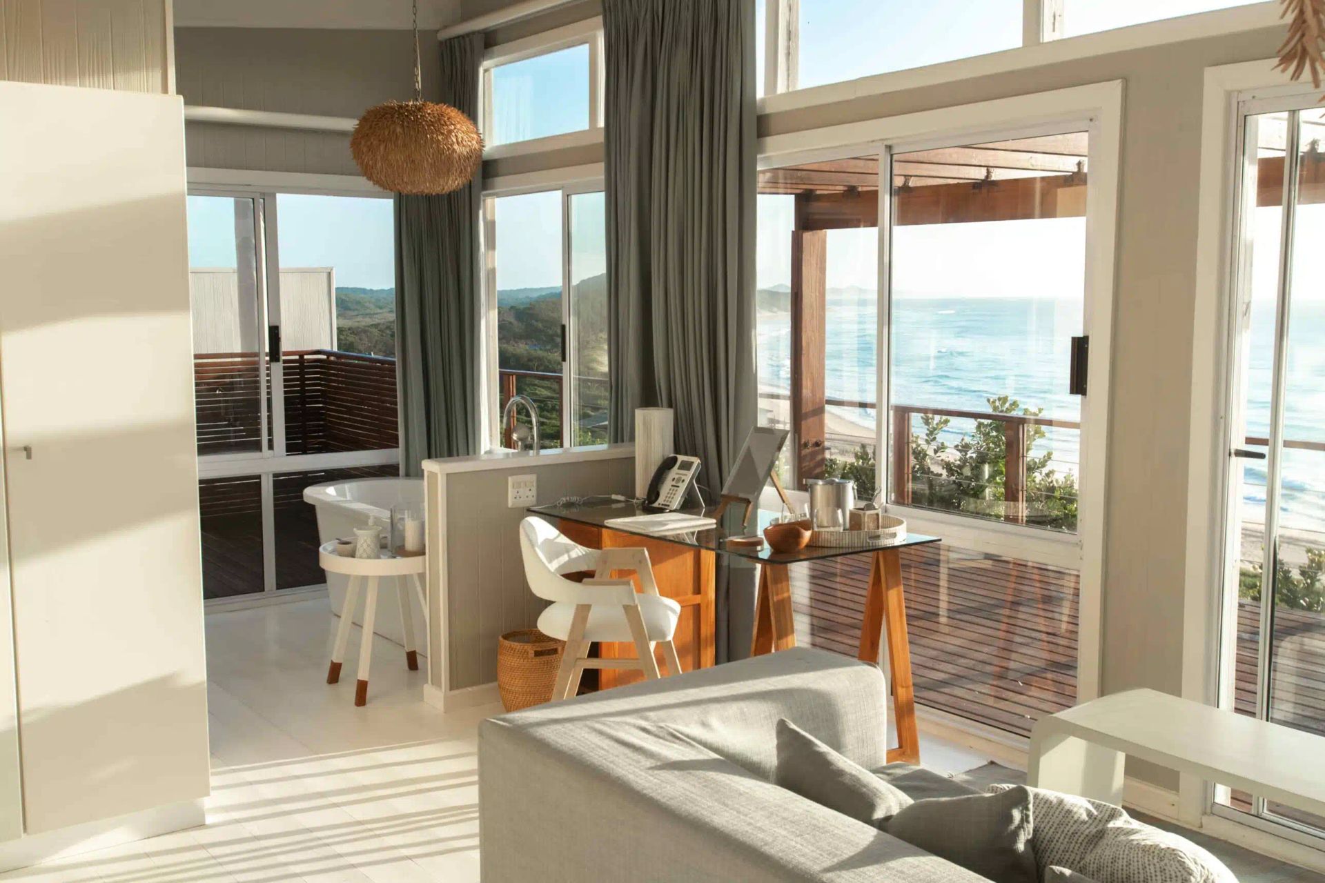 Bright seaside dining room with sunlit table, white chairs, and ocean view through large windows