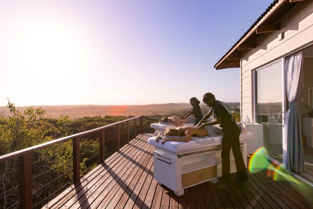 Two people on an outdoor massage table on a sunlit deck overlooking a field