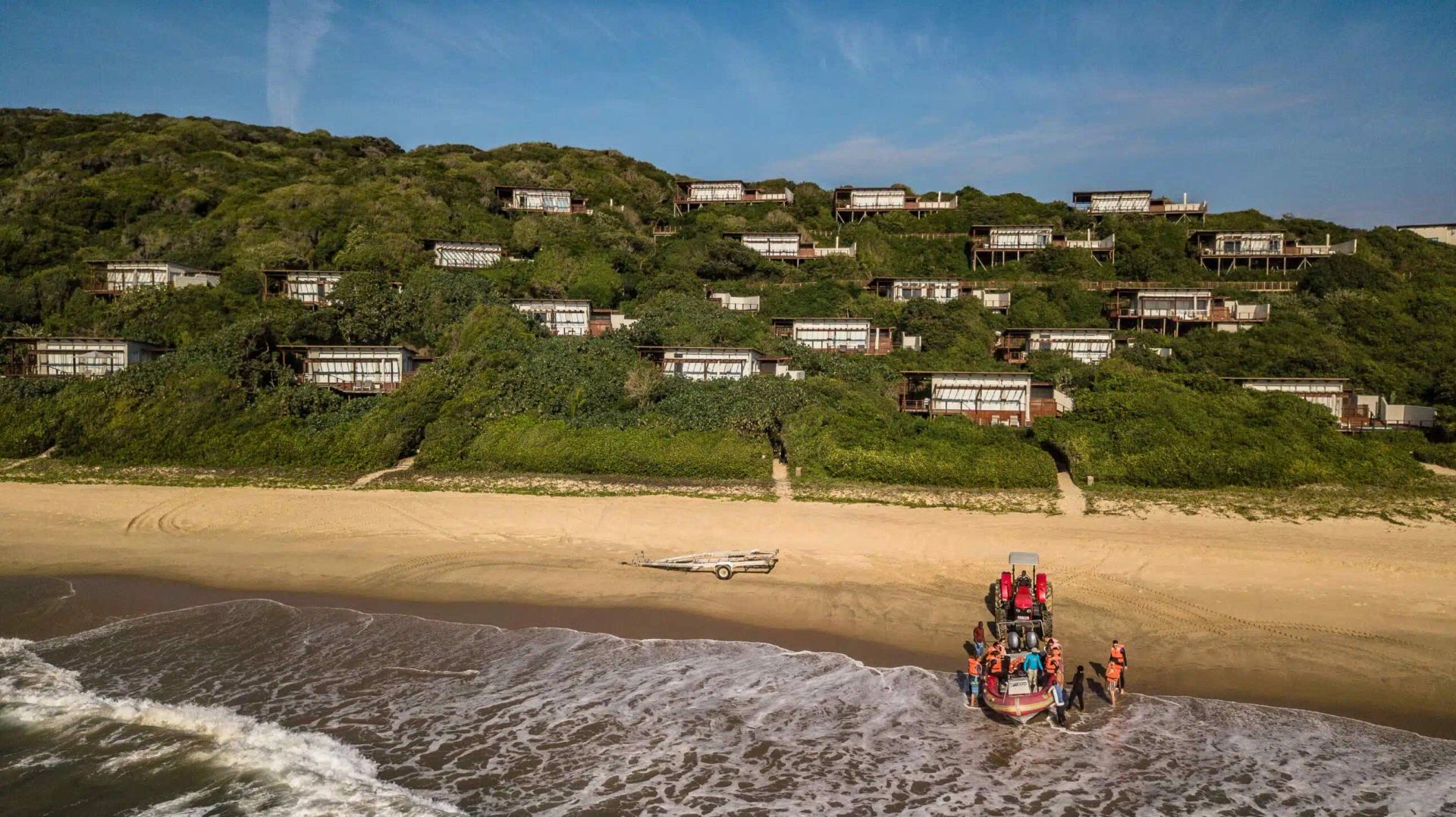 A boat with people lands on a sandy beach below a hillside covered in lush green vegetation and scattered houses.