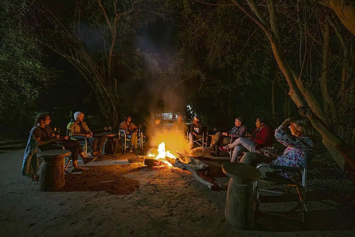 A group of people sits in camp chairs around a glowing campfire at night in a wooded area.