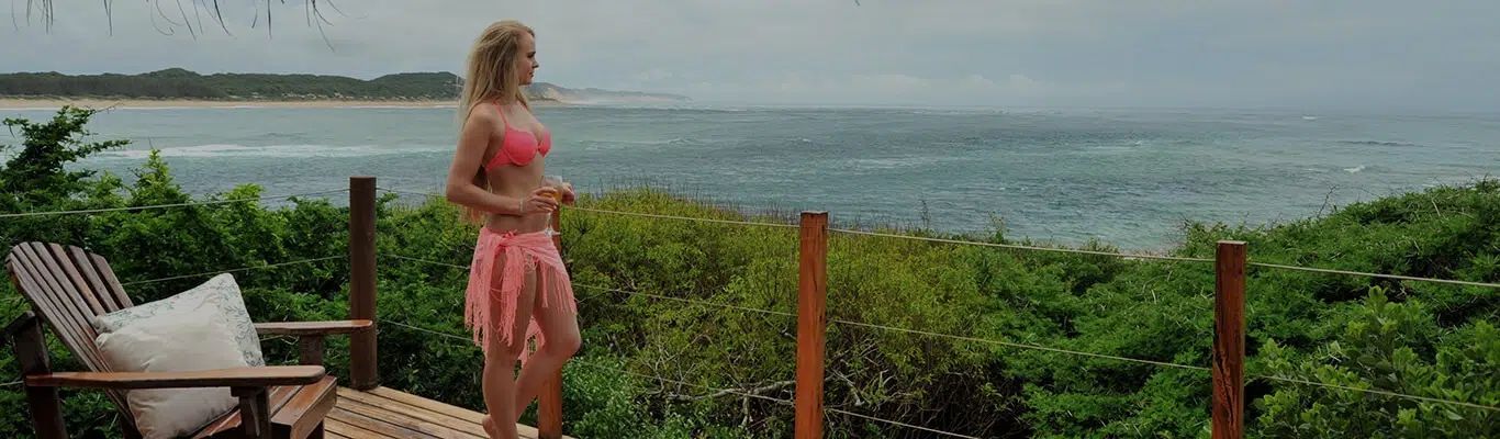 Woman in a pink dress standing on a balcony, looking out over the ocean and green coastline.