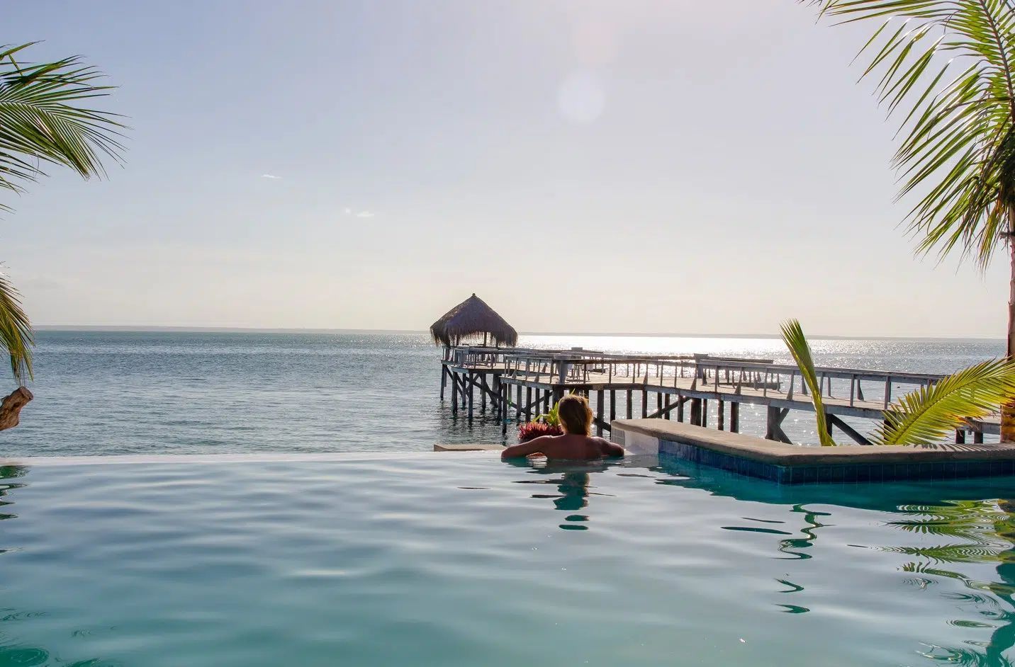 Infinity pool overlooking the ocean with a wooden pier and thatched hut at sunset