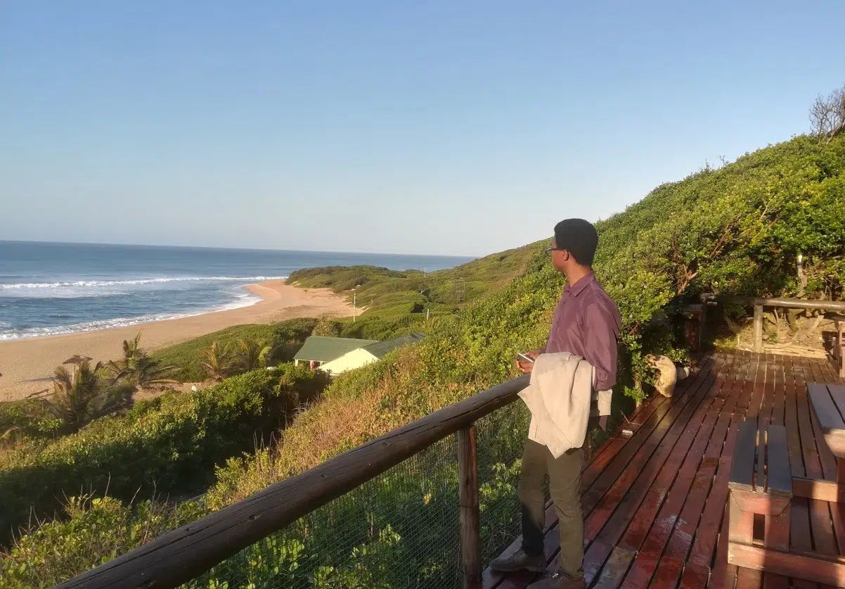 A person stands on a wooden deck overlooking a sandy beach and ocean, with lush green coastal vegetation in the background.
