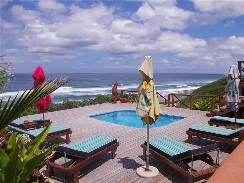 Wooden pool deck overlooking the ocean with lounge chairs, closed umbrellas, and a small pool under a cloudy blue sky.