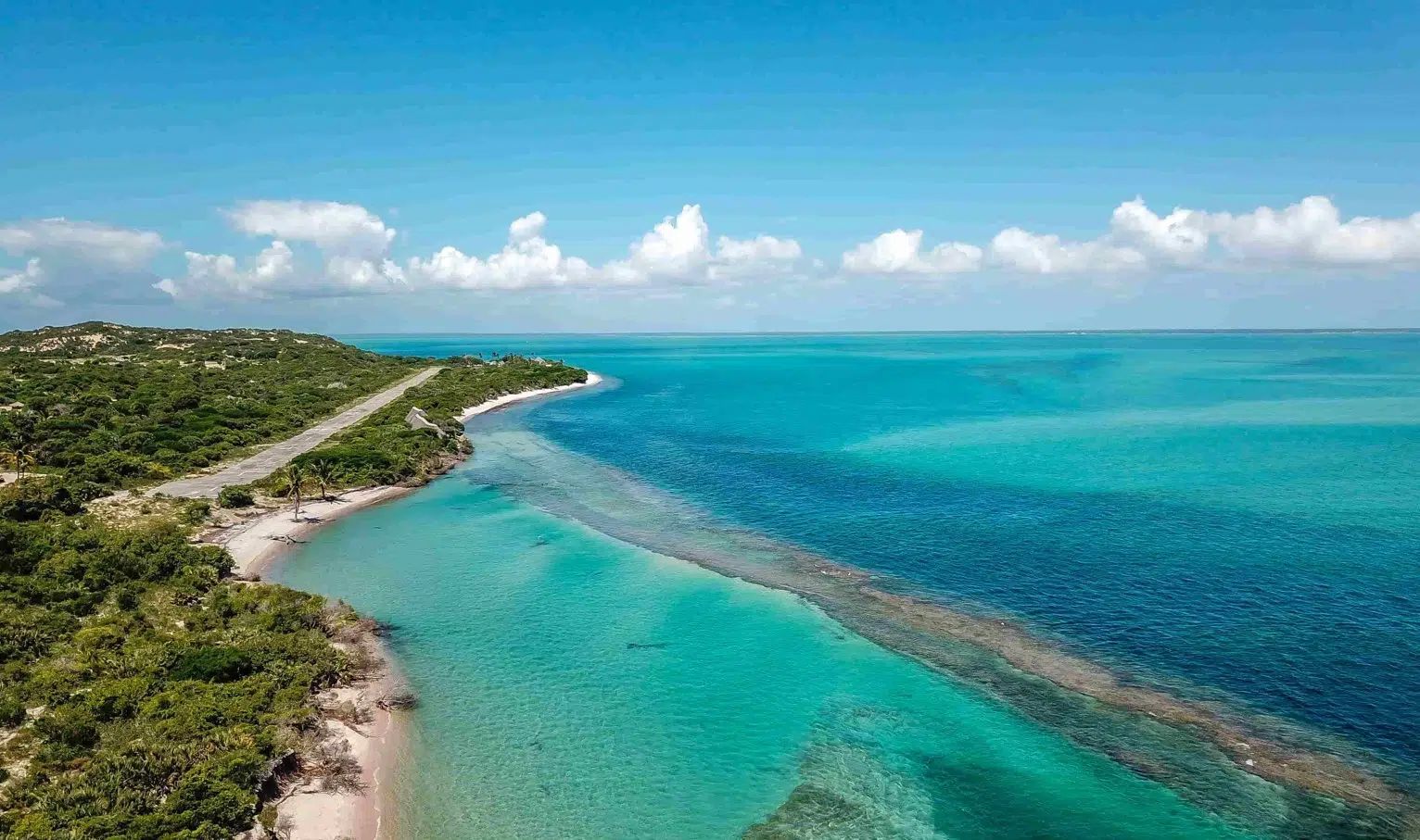 Aerial view of a turquoise tropical coastline with a winding road and blue ocean under a cloudy sky