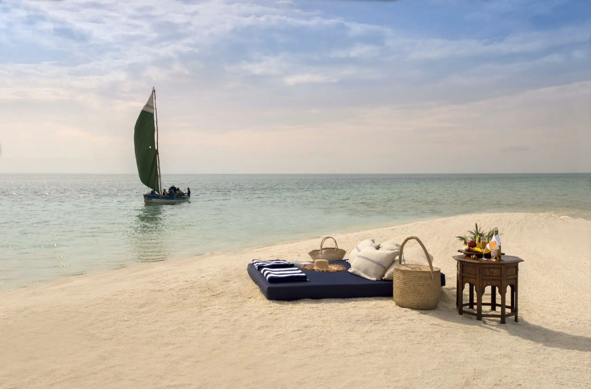 Beach lounge setup with blue cushions, wicker table, and sailboat on calm tropical water