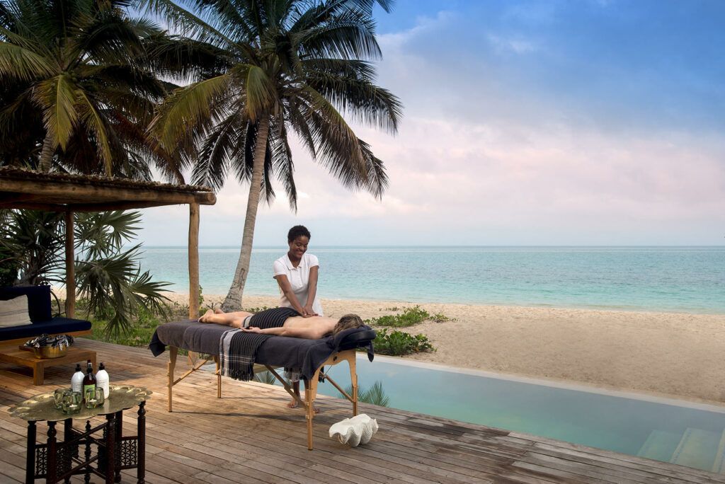 Massage table by a poolside beach cabana at sunset, with palm trees and ocean beyond.