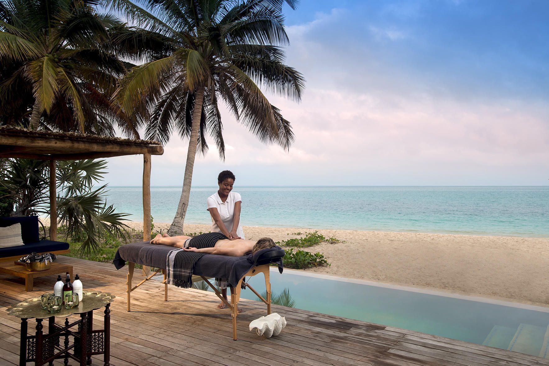 Massage table on a tropical beach beside a pool, with palm trees and ocean at sunset.