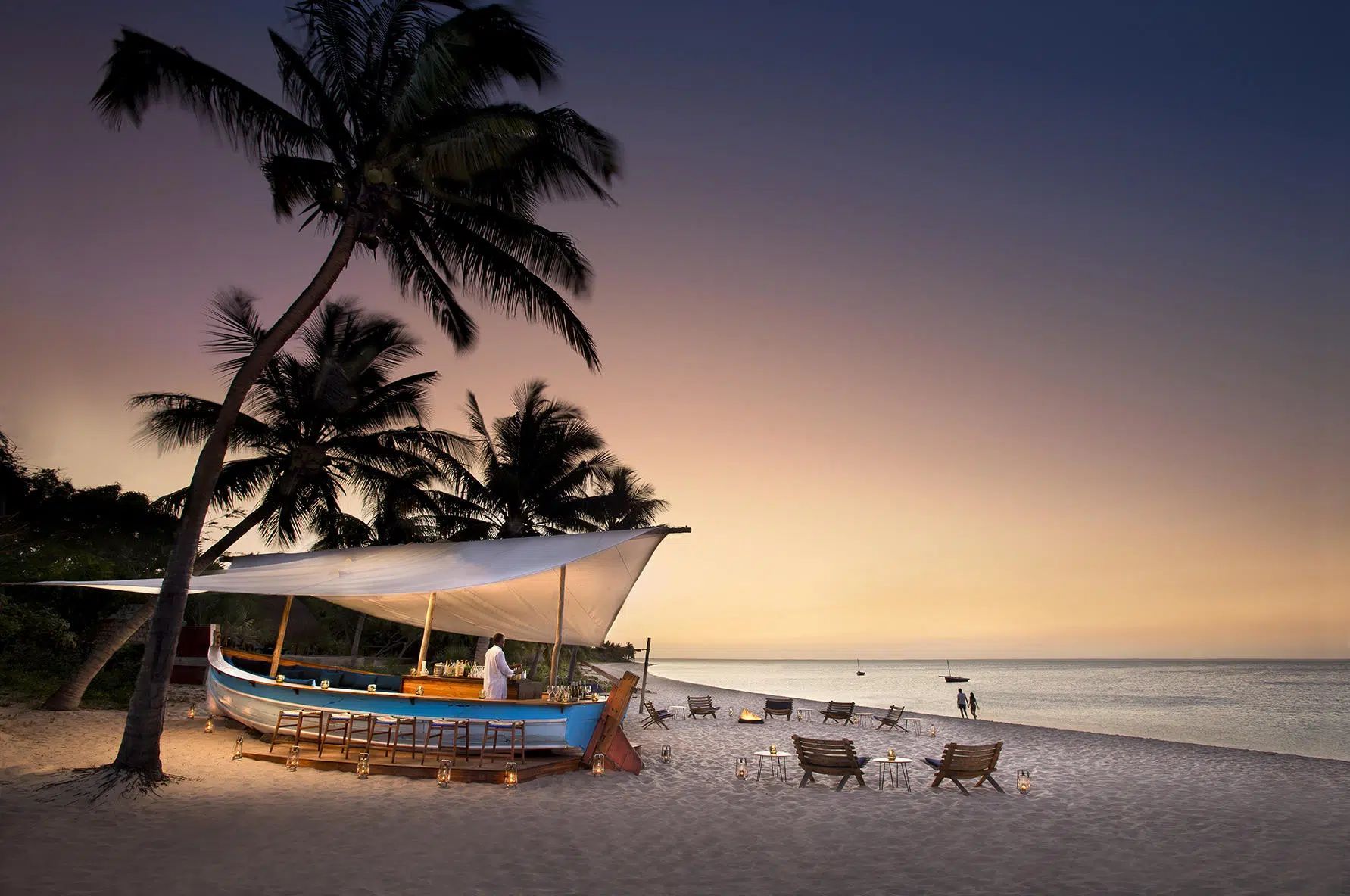 Beach at sunset with palm trees, a small boat, and chairs on the shore