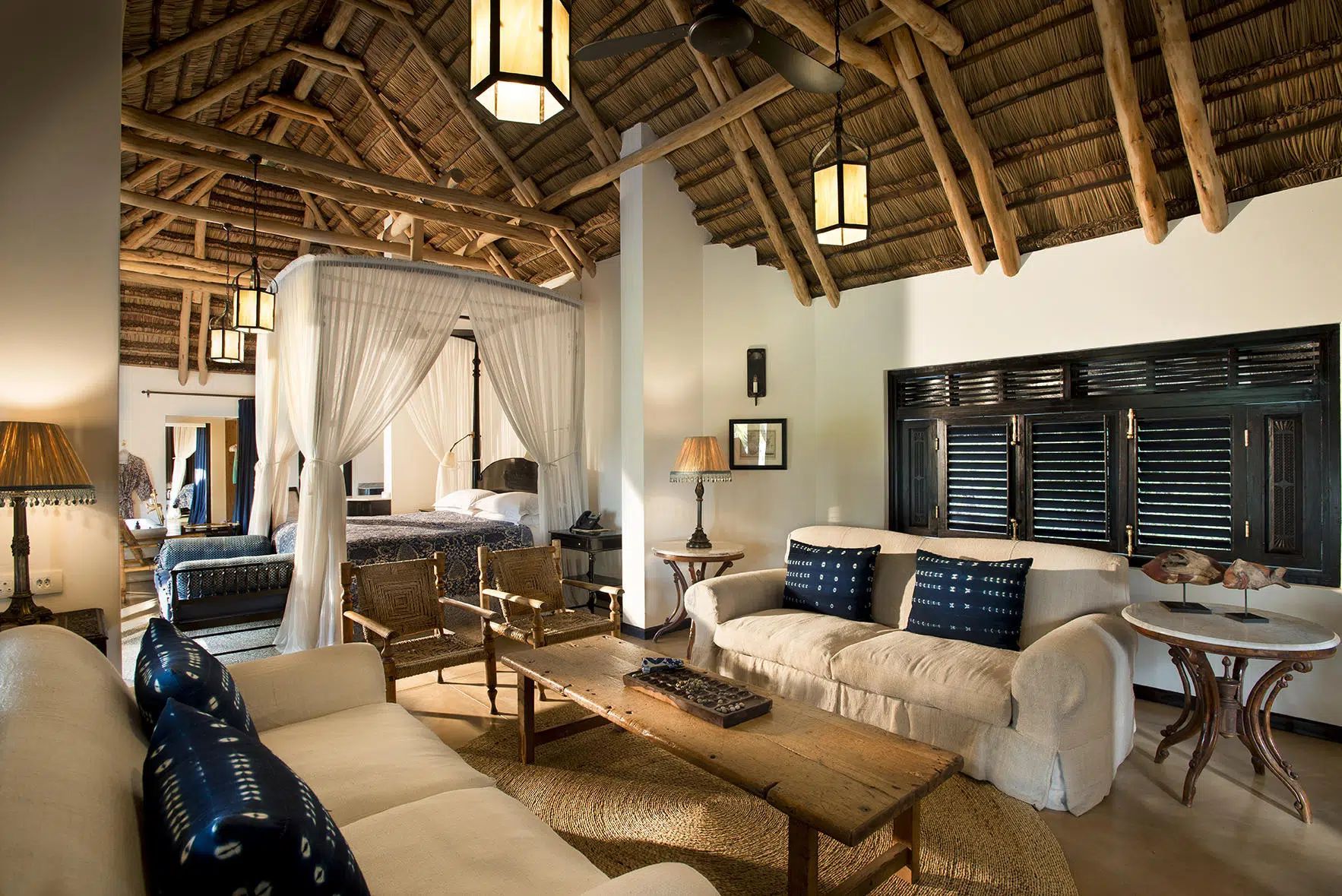 Rustic bedroom with canopy bed, woven ceiling, white walls, blue and beige seating, and warm lantern lighting