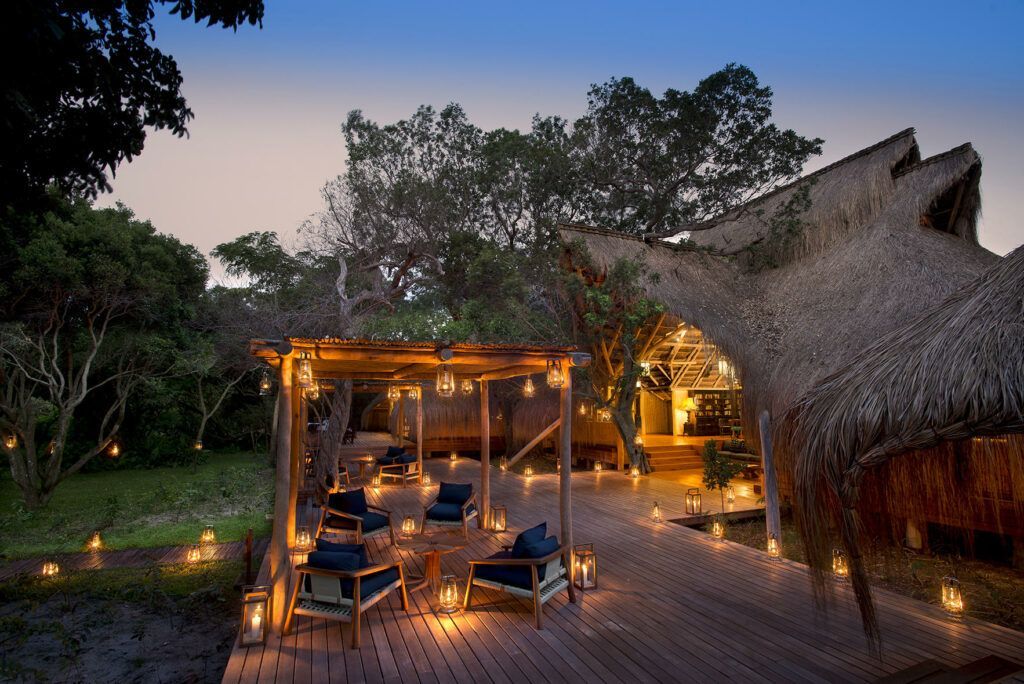Tropical resort deck at dusk with lit lounge seating and a thatched-roof pavilion