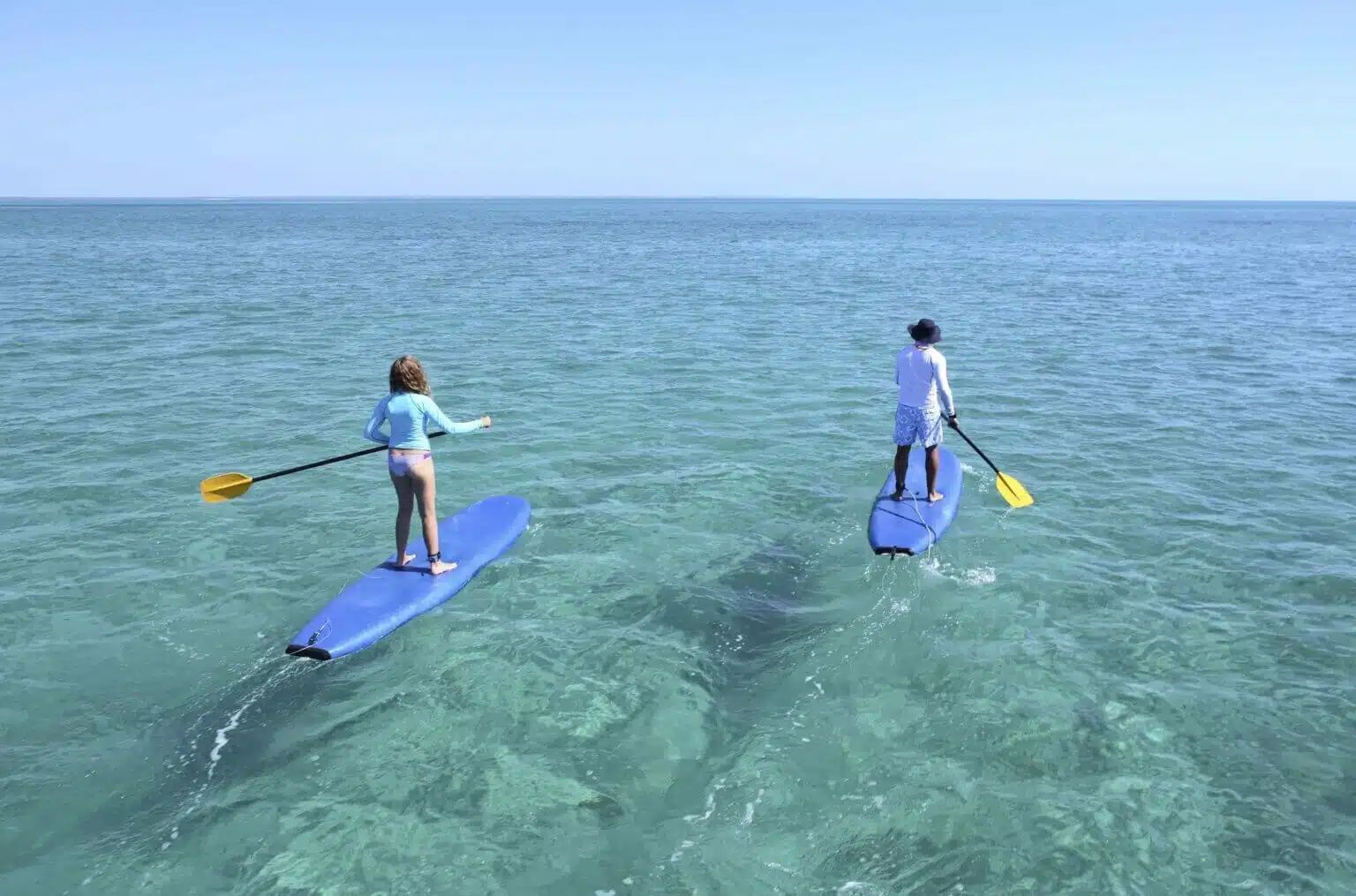 Two people paddleboard on clear, calm water under a bright blue sky.
