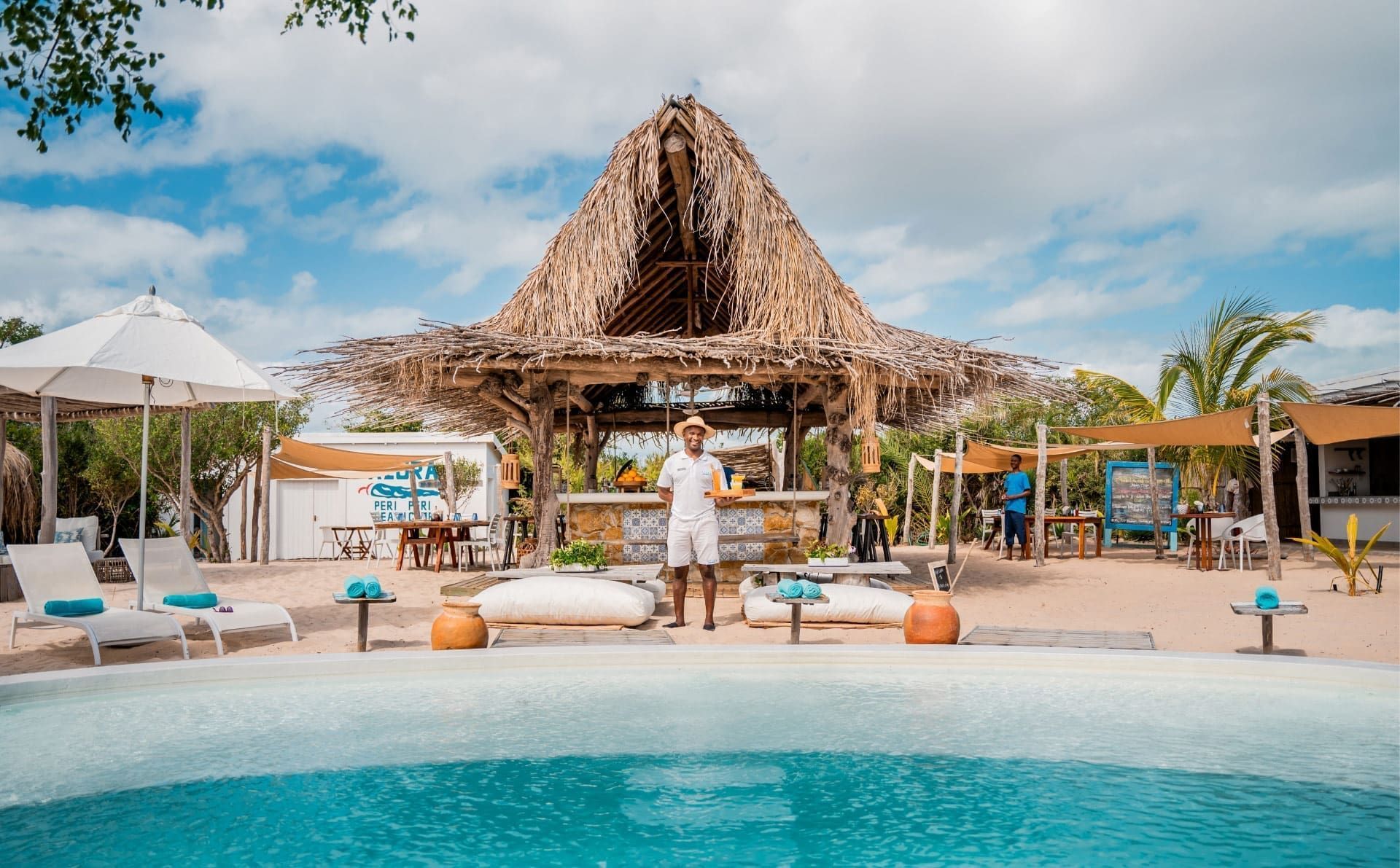 Tropical poolside lounge with thatched cabana, white umbrellas, and turquoise water under a bright sky
