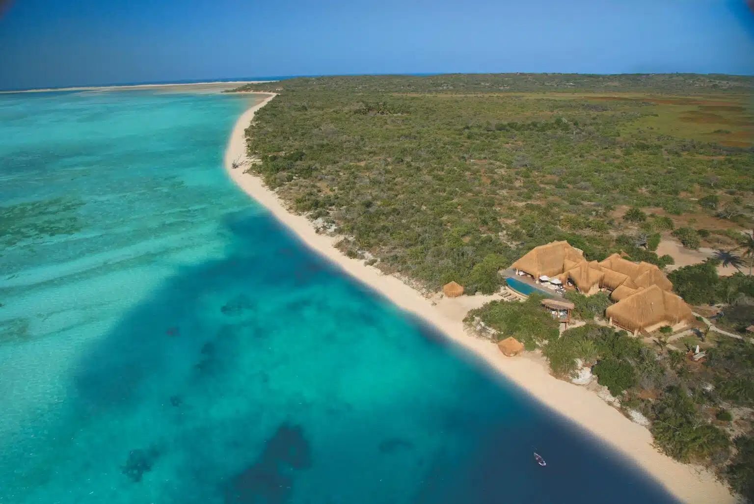 Aerial view of a luxury thatched-roof resort nestled between a lush green forest and a turquoise tropical beach.