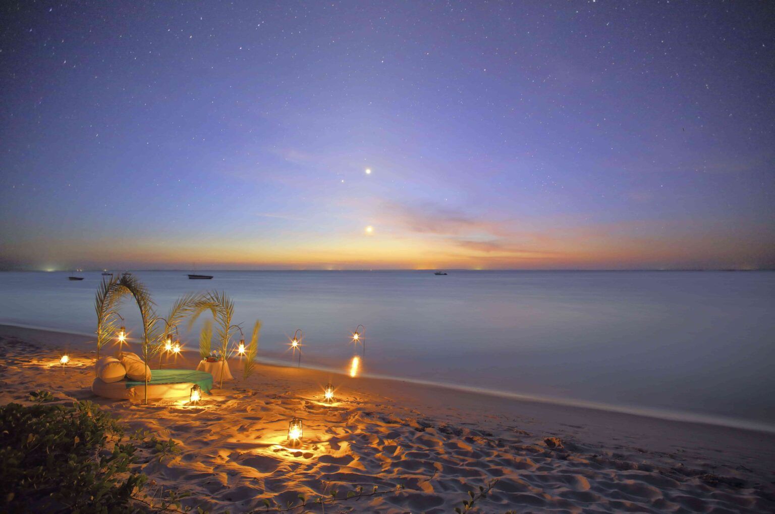 A candlelit romantic setup with floral arches on a sandy beach at twilight, under a star-filled sky.