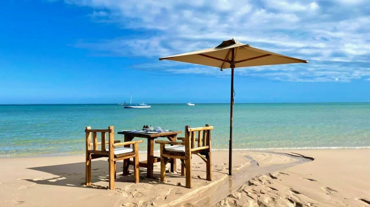 Beachside table and chairs under a sun umbrella on sandy shore, with calm blue sea and sailboat in the distance.
