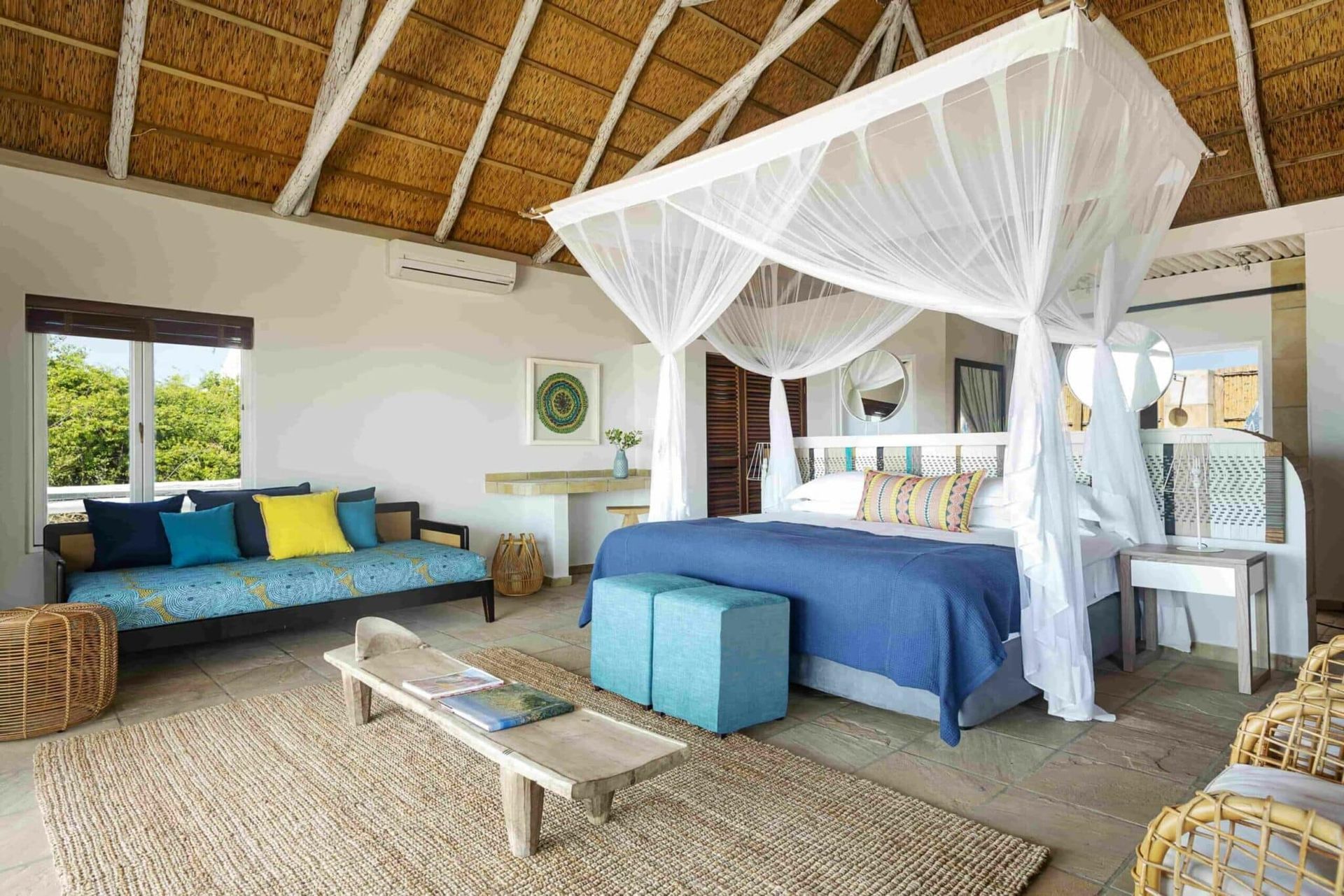 Tropical bedroom with a blue bed, white canopy netting, and a sitting area under a thatched roof