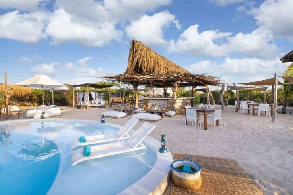Beachside pool with white lounge chairs, thatched cabanas, and tables under a sunny sky