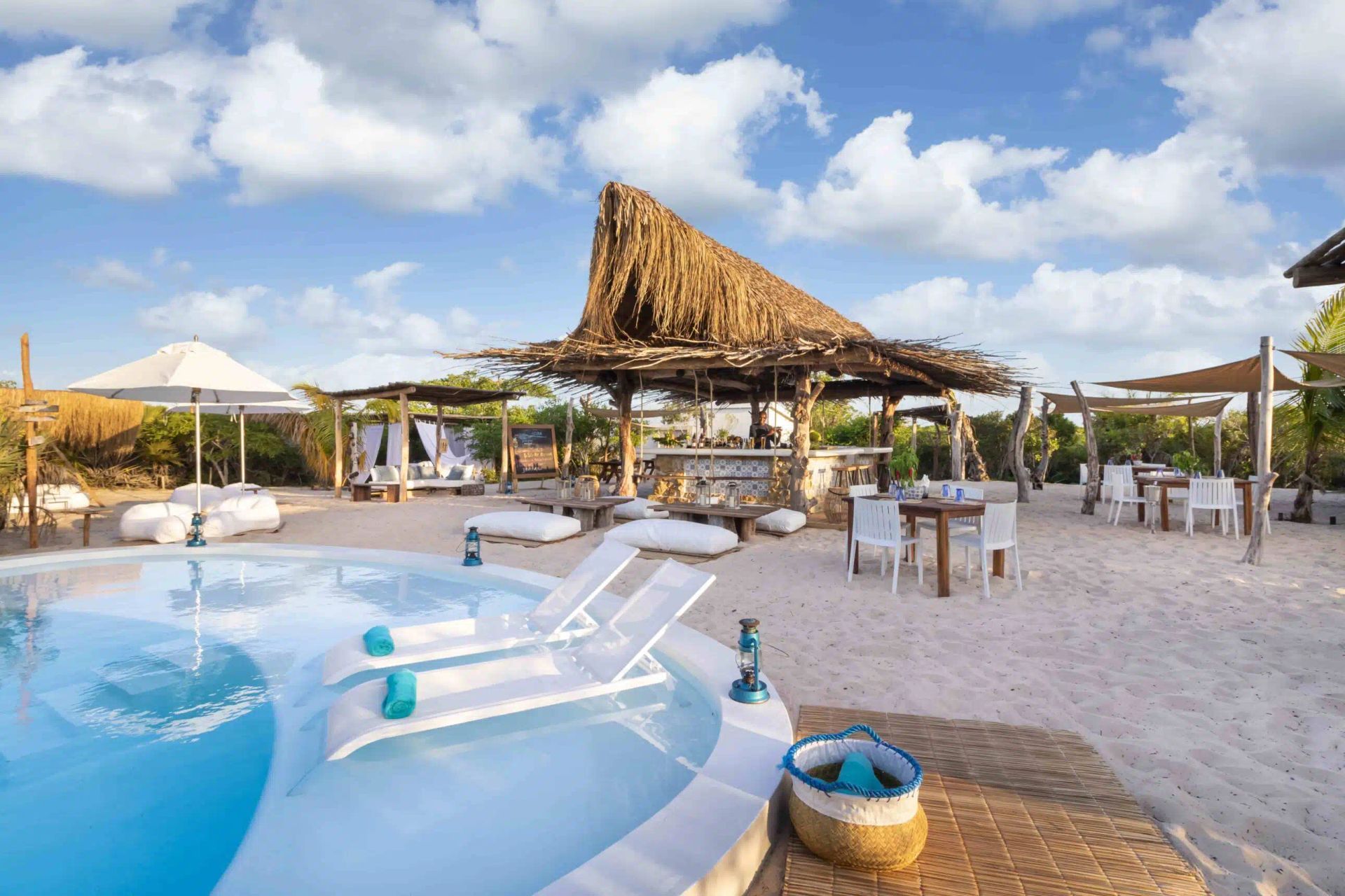 A sandy beach bar and pool area with white lounge chairs, straw-roofed structures, tables, and a bright blue sky.