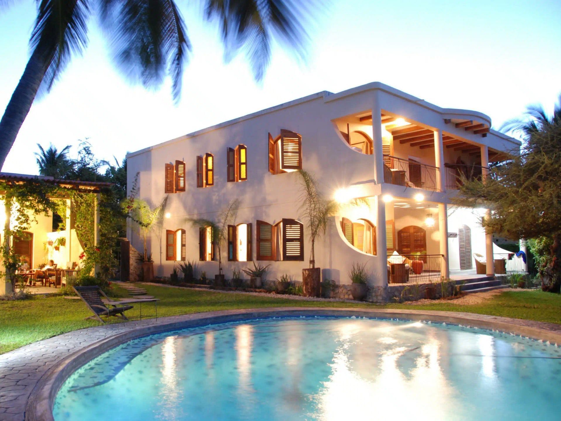 Luxury villa with lit windows beside a curved pool at dusk, framed by palm trees.