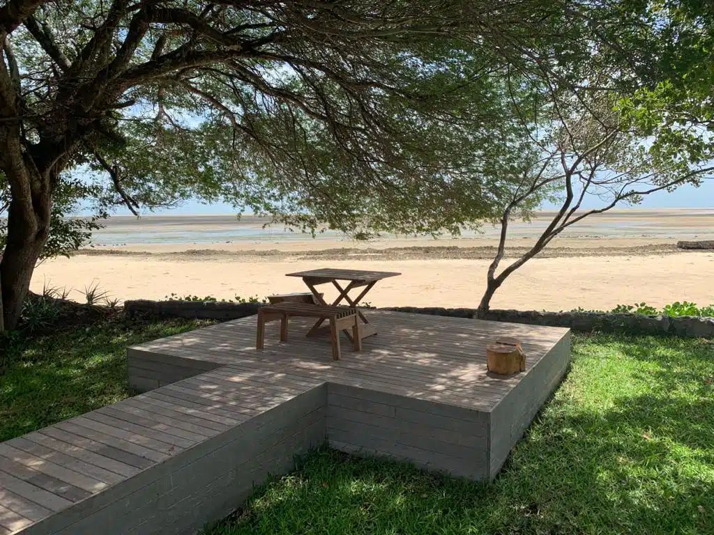 Shaded wooden deck with table and chairs overlooking a sandy beach and calm water