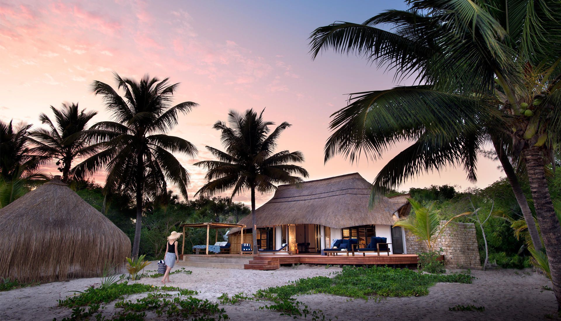 Tropical beach resort at sunset with palm trees, thatched hut, and lit villa by the sand.