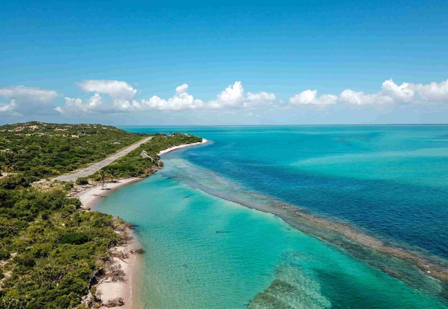 Aerial view of a turquoise tropical shoreline with green hills, white clouds, and a long reef line in the sea