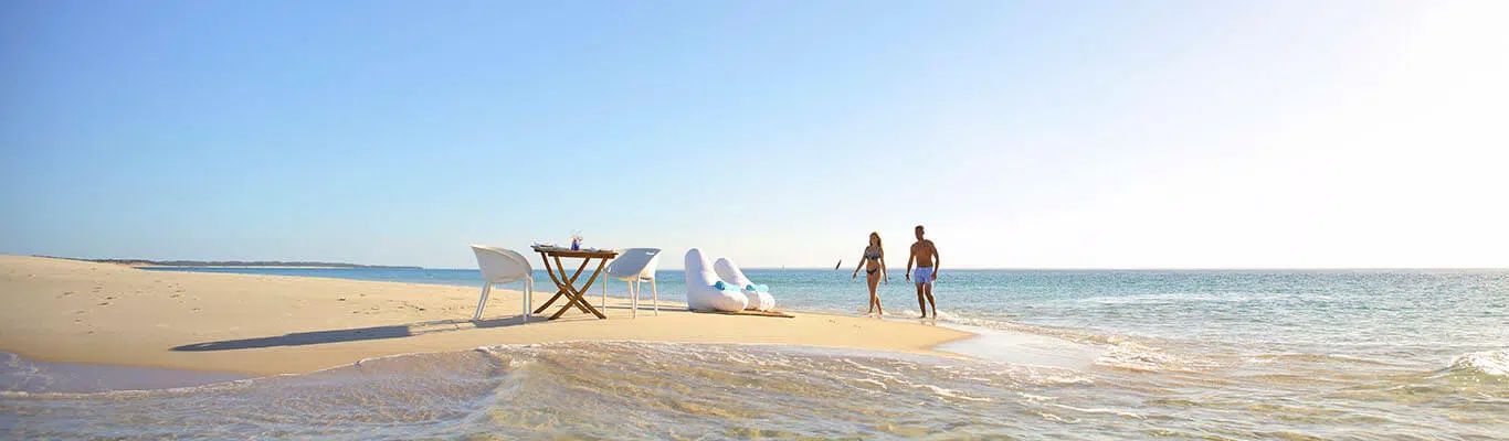 People walking on a sunny beach near the ocean, with a lounge chair in the sand.