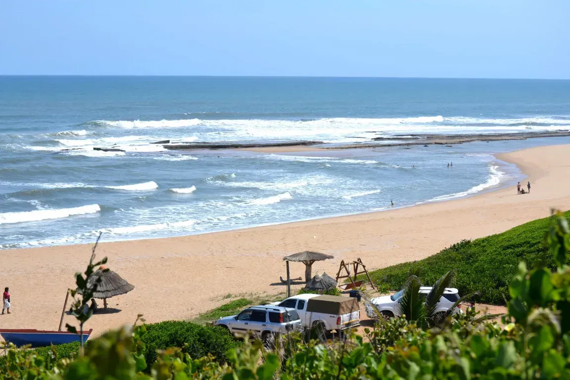 A sunny beach with light sand, crashing waves, thatched umbrellas, parked vehicles, and green coastal vegetation.