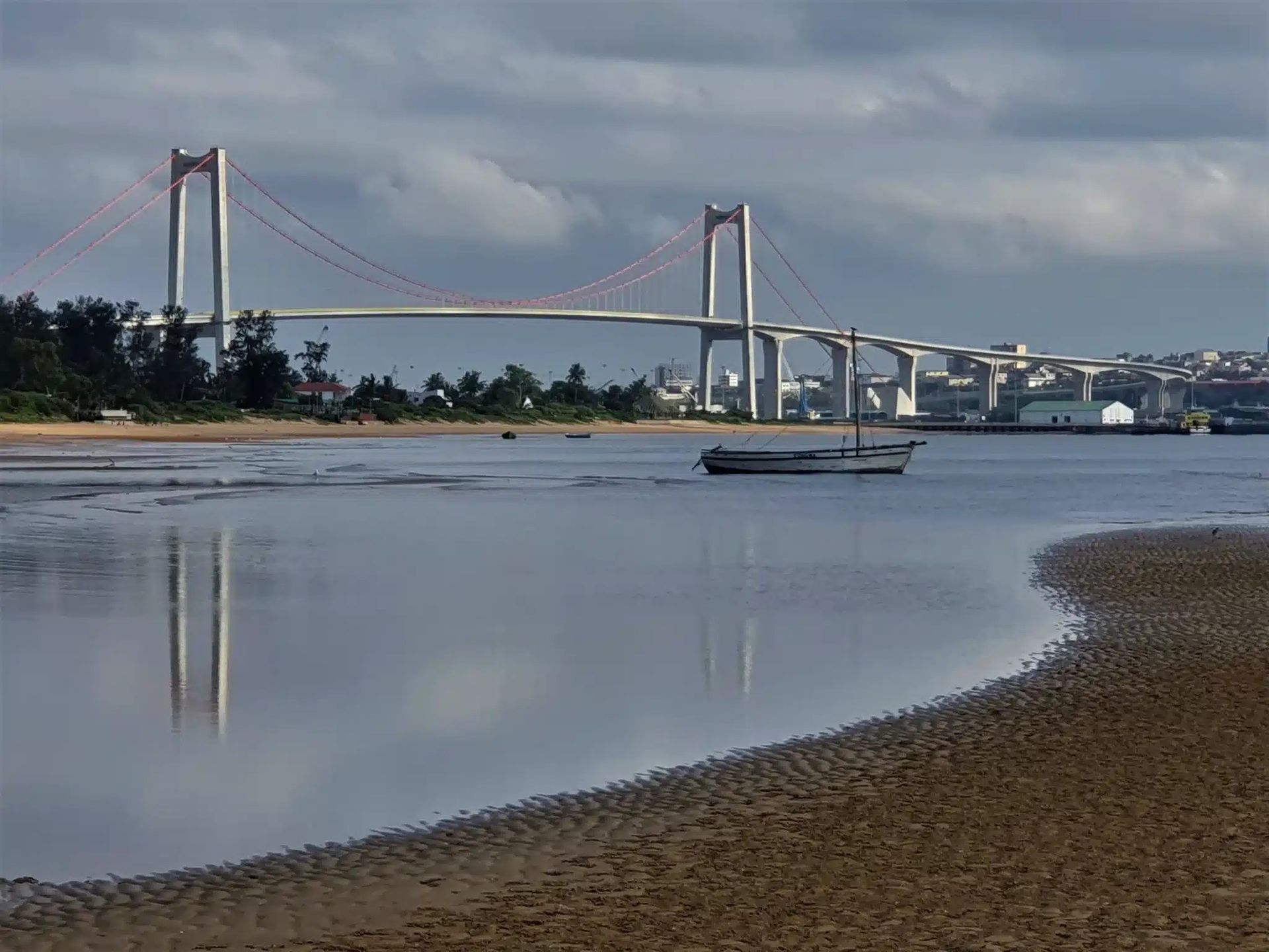 Suspension bridge over calm water with a small boat, cloudy sky, and shoreline at low tide