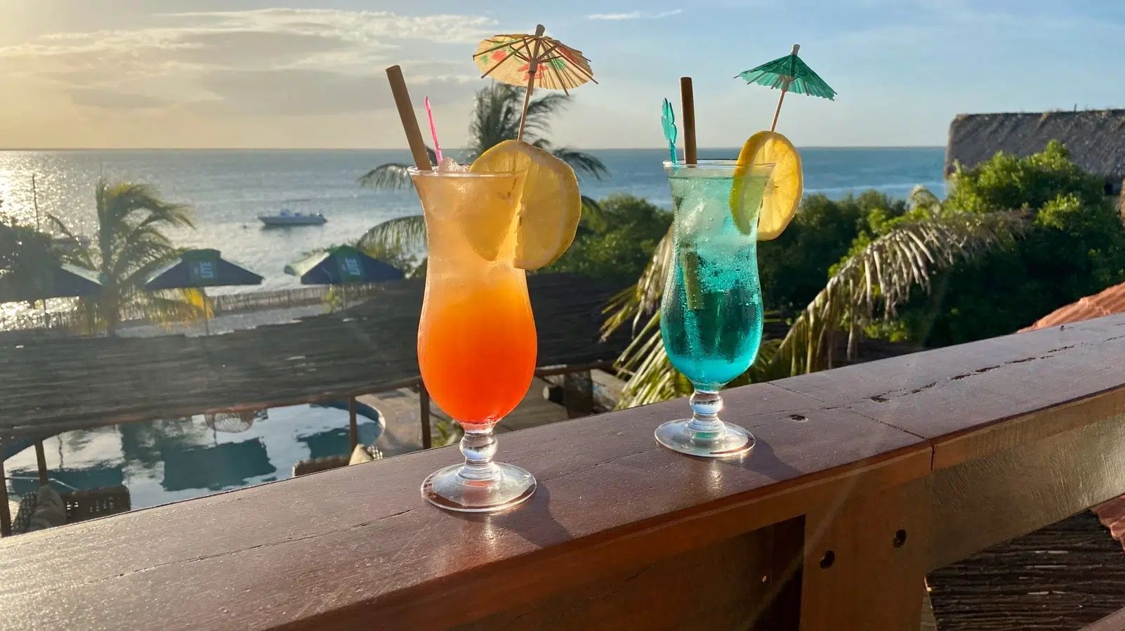 Two colorful cocktails on a railing overlooking a beach at sunset