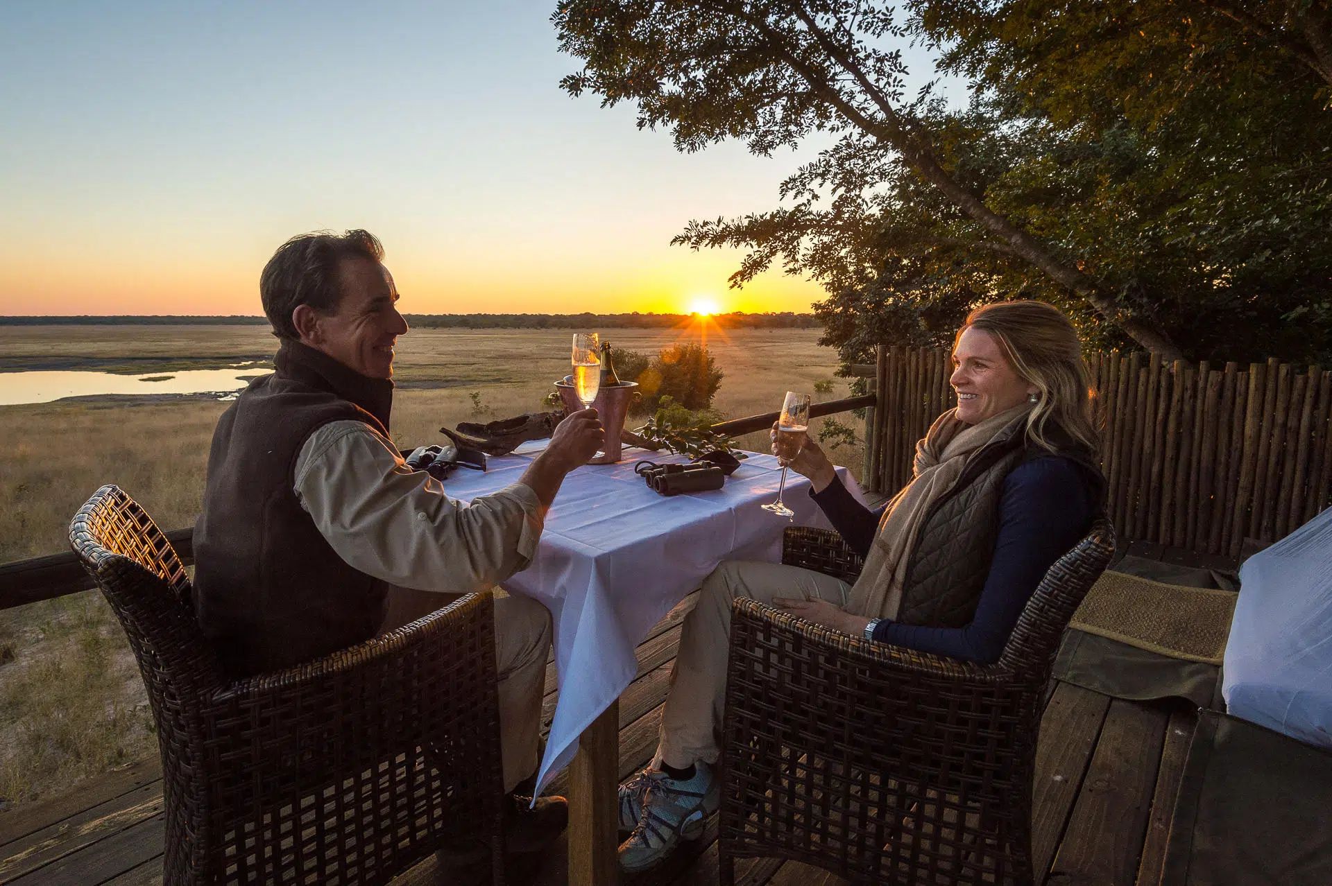 Two people dining at sunset by a waterfront, raising glasses across a candlelit table