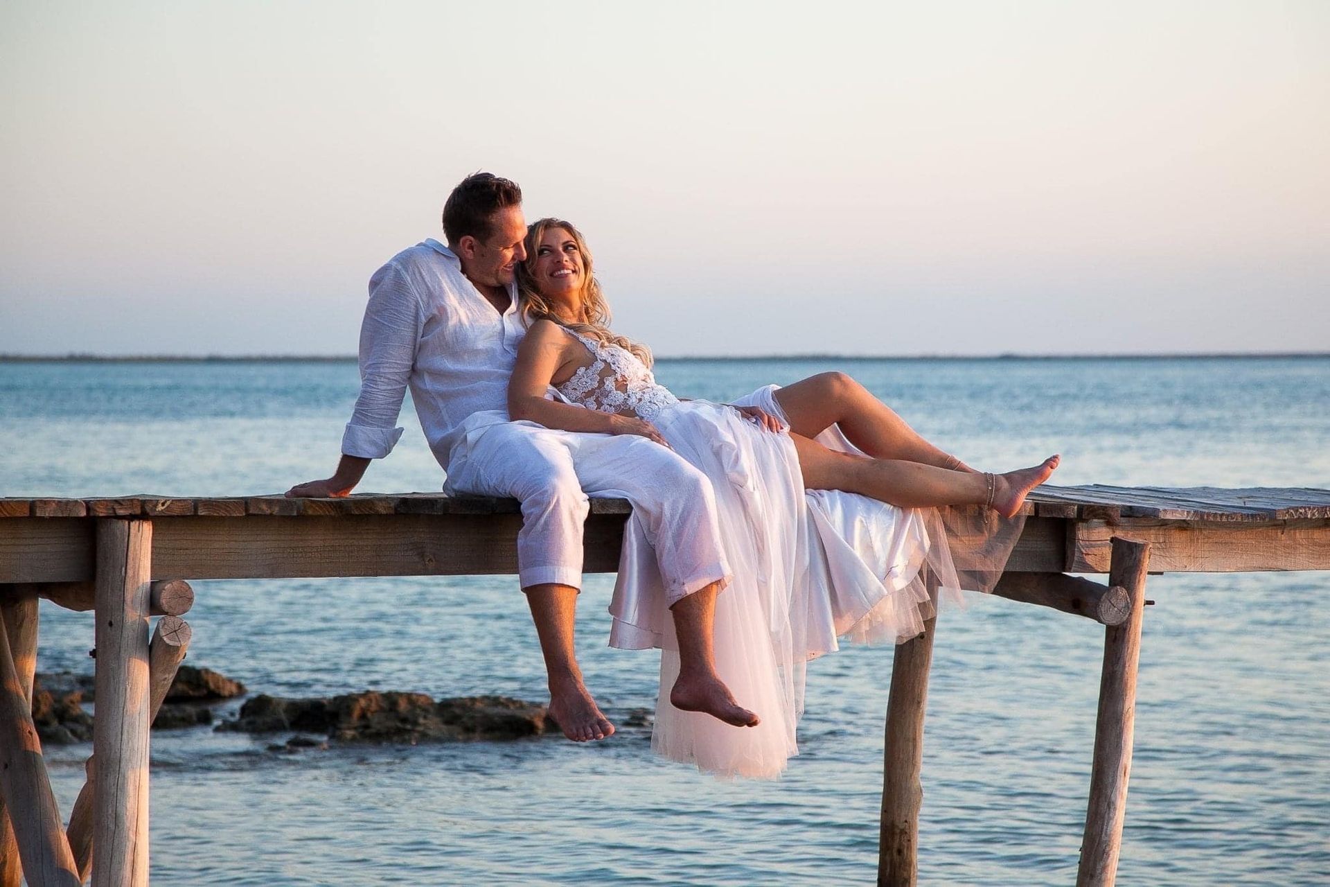Couple in white clothing sitting on a pier at sunset by the sea, feet dangling above the water.