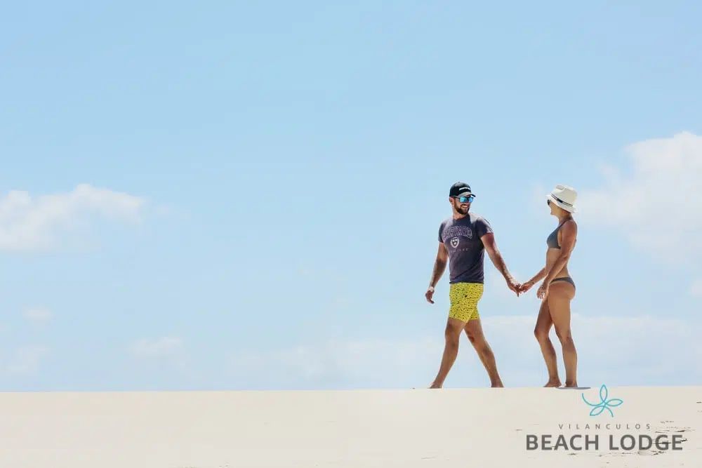 A couple walks hand-in-hand across a vast, bright sand dune under a clear blue sky, with a beach lodge logo below.