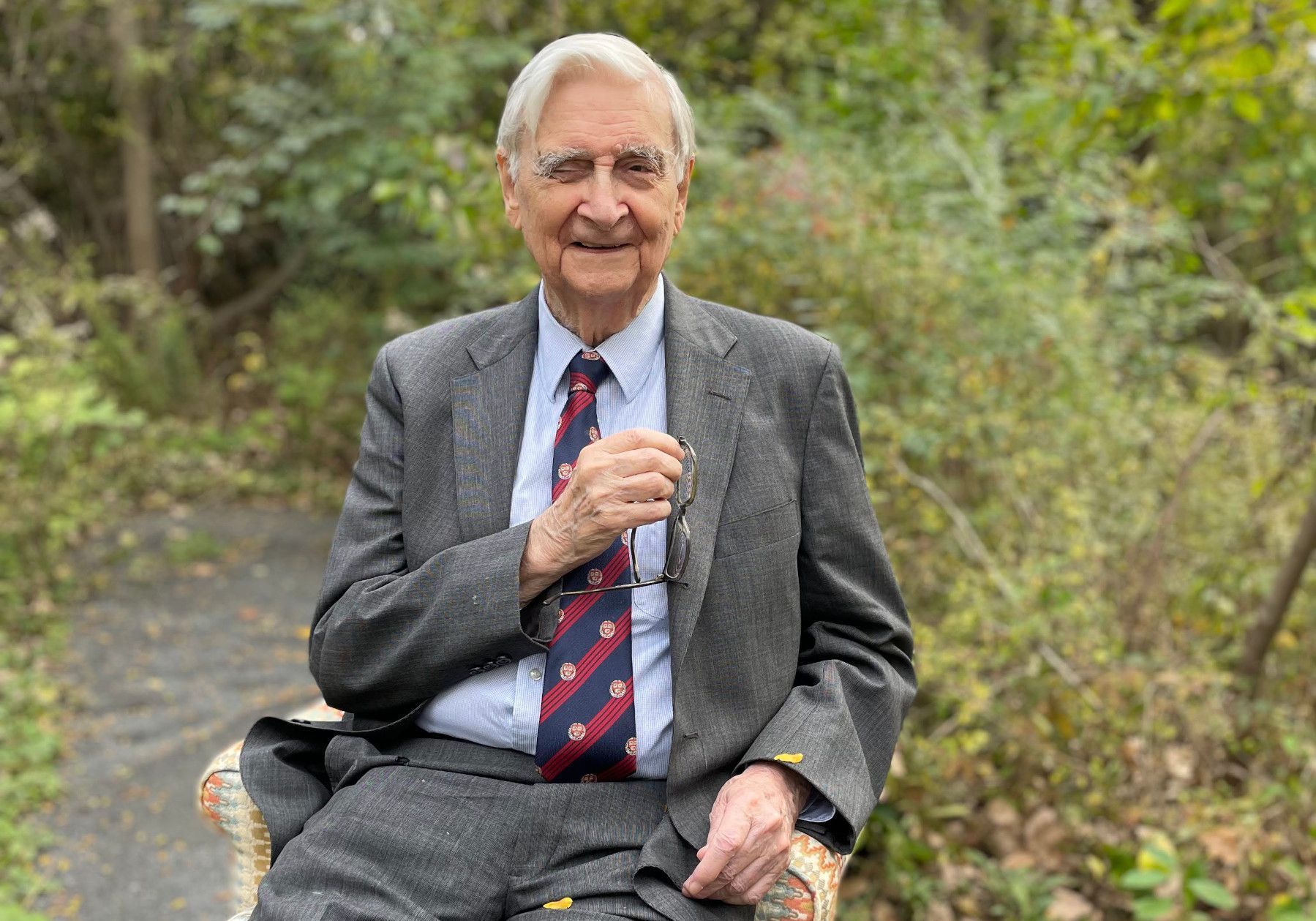 Elderly man in a gray suit and red tie sitting outdoors, smiling and holding a cane.