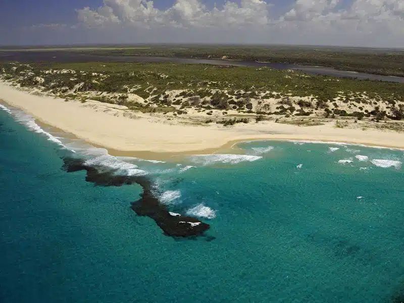 Aerial view of a sandy beach meeting turquoise water, with a dark rock formation and green vegetation along the coast.
